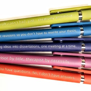 Op de afbeelding: Een verzameling kleurrijke pennen in groen, blauw, paars, roze en rood. Elke pen heeft een andere humoristische zin erop gedrukt, zoals "Je concept was niet slecht, gewoon... creatief ambitieus."