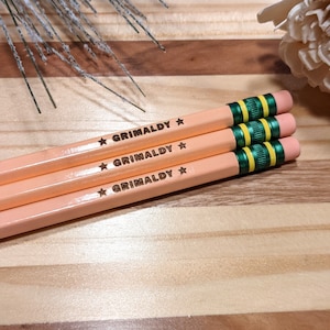 May include: Three peach-coloured pencils with the word "GRIMALDY" printed on them in black. Each pencil has a green and yellow striped band near the eraser end. The erasers are pink. The pencils are arranged on a wooden surface.