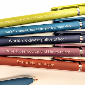 Puede incluir: Una colección de bolígrafos coloridos con frases humorísticas impresas. Los bolígrafos son de color verde, azul, morado y rojo. El texto en los bolígrafos incluye frases como "The police never think it's as funny as you do" y "Definitely not a cop".