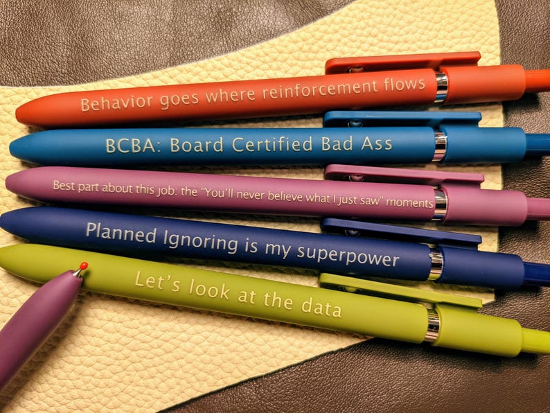 May include: Six colorful pens with different motivational quotes printed on them. The quotes are: "Behavior goes where reinforcement flows", "BCBA: Board Certified Bad Ass", "Best part about this job, the "You'll never believe what I just saw" moments", "Planned Ignoring is my superpower", and "Let's look at the data".