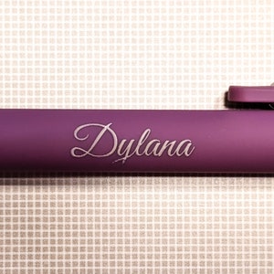 May include: A close-up of a purple pen with the name "Dylana" elegantly engraved in a cursive font. The pen's body has a matte finish, and the background is a textured, off-white surface.