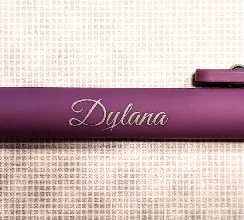 May include: A close-up of a purple pen with the name "Dylana" elegantly engraved in a cursive font. The pen's body has a matte finish, and the background is a textured, off-white surface.