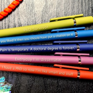 May include: Five colourful pens with humorous text about PhDs. The pens are green, blue, dark blue, pink, and orange. The text on the pens reads: "PhD Survivor", "If you liked it then you should have put a citation on it", "Pain is temporary. A doctoral degree is forever.", "Everyday I'm dissertatin'", and "I have a PhD so now I'm like smart and stuff".