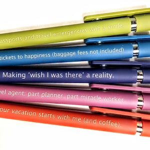 May include: A selection of colourful pens with witty travel-themed quotes in white. The pens are in shades of green, blue, purple, pink, and red. Each pen has a silver clip and tip.