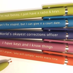 Puede incluir: Una colección de bolígrafos coloridos con frases humorísticas impresas. Los bolígrafos son en tonos verdes, azules, morados, rosas y naranjas. Cada bolígrafo tiene un clip y una punta a juego. El texto en los bolígrafos incluye frases como "No soy mandona" y "No puedo arreglar la estupidez".