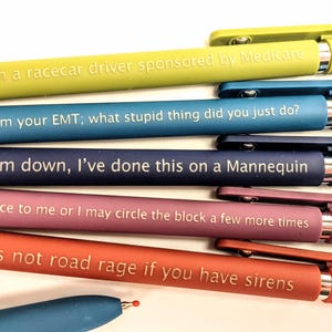 May include: A collection of colorful pens with humorous phrases printed on them. The pens are in shades of yellow, blue, navy, pink, and red. The text includes phrases like "I'm a racecar driver sponsored by Medicare" and "It's not road rage if you have sirens."