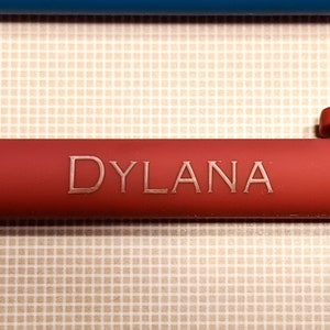 May include: Close-up of a red pen with the name "DYLANA" engraved in a lighter color. The pen is set against a patterned background with a blue object in the background.