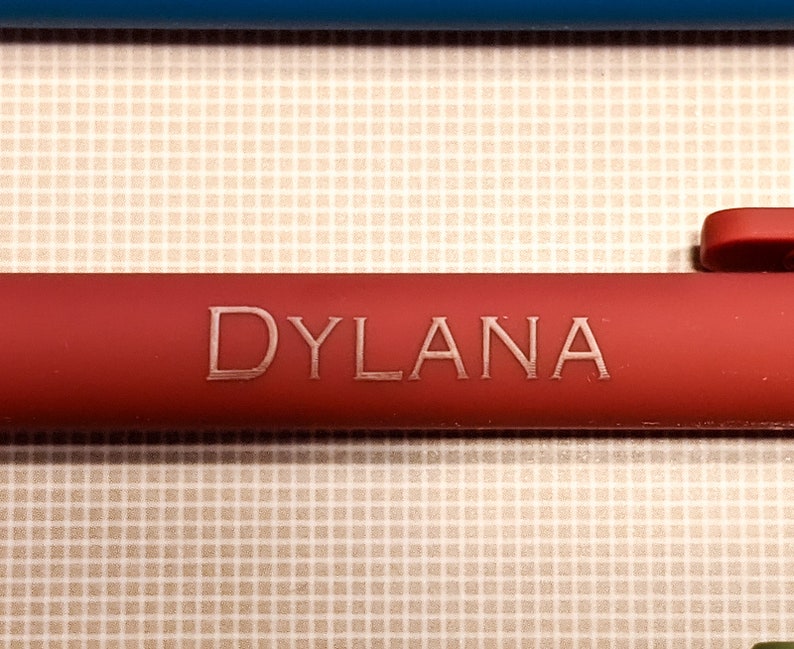 May include: Close-up of a red pen with the name "DYLANA" engraved in a lighter color. The pen is set against a patterned background with a blue object in the background.
