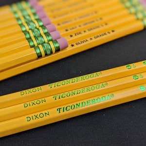 May include: Close-up of yellow Dixon Ticonderoga pencils with pink erasers and green accents. The pencils are labeled with the brand name and the word "SOFT". Some pencils are labeled with names and "GRADE 5".