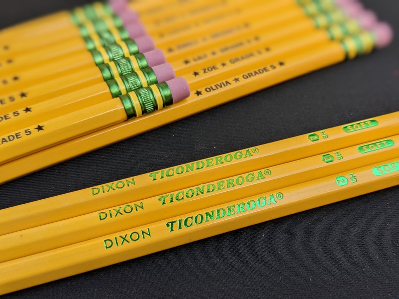 May include: Close-up of yellow Dixon Ticonderoga pencils with pink erasers and green accents. The pencils are labeled with the brand name and the word "SOFT". Some pencils are labeled with names and "GRADE 5".