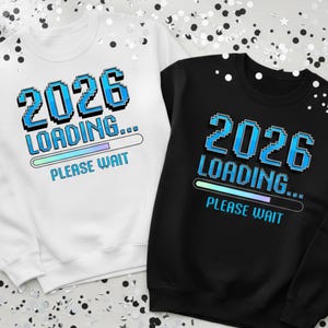 May include: Two sweatshirts, one white and one black, each featuring the text "2026 LOADING... PLEASE WAIT" in pixelated blue font. The loading bar is a gradient of blue and green. The sweatshirts are surrounded by confetti.