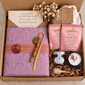 Open book gift box featuring a beautifully wrapped book in pink and lavender patterned paper, handcrafted beaded bookmark with charm, wax seal, affirmation card, milk tea, mini honey jar, wooden honey dipper, candle, and sweet treat. Thoughtfully curated reading and self-care gift by Gratefully Created.