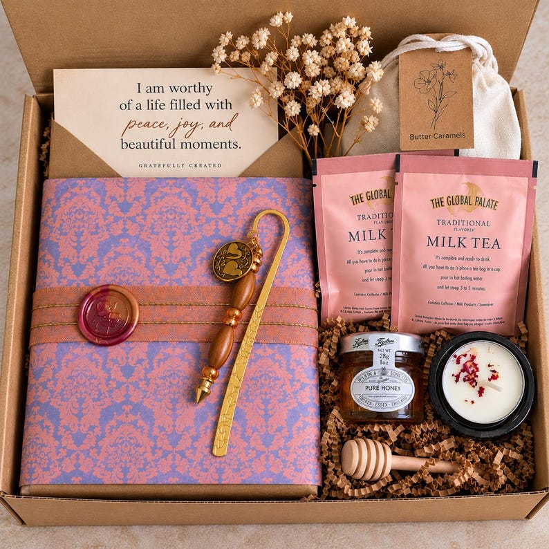 Open book gift box featuring a beautifully wrapped book in pink and lavender patterned paper, handcrafted beaded bookmark with charm, wax seal, affirmation card, milk tea, mini honey jar, wooden honey dipper, candle, and sweet treat. Thoughtfully curated reading and self-care gift by Gratefully Created.
