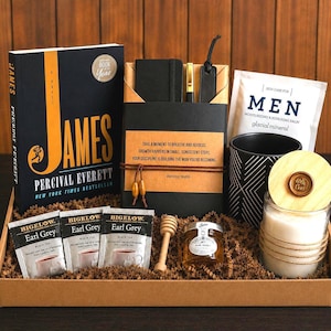 May include: A gift box containing a James novel by Percival Everett, a black notebook with a gold pen, a Men moisturizing balm packet, a black patterned mug, a candle with a wooden lid, three Bigelow Earl Grey tea bags, and a honey dipper.