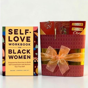 May include: A gift set featuring a book titled "Self-Love Workbook for Black Women" and a burgundy box with gold ribbon. The box contains tea, a pen, and a decorative flower. The book's cover is yellow and burgundy.