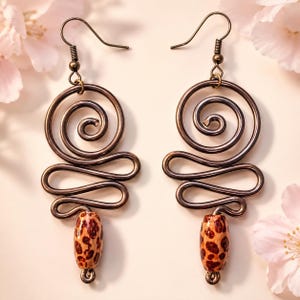 May include: A pair of dark bronze-toned metal earrings featuring a wire-wrapped spiral design at the top, a wavy wire section in the middle, and a single cylindrical bead with a brown and tan leopard print pattern hanging at the bottom.