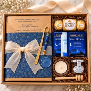 A gift box containing a blue-wrapped journal with a white ribbon, a gold-toned metal bookmark with blue beads, a gold pen, two Ferrero Rocher chocolates, two Bigelow Sweet Dreams tea bags, a small candle, and a jar of honey with a dipper.