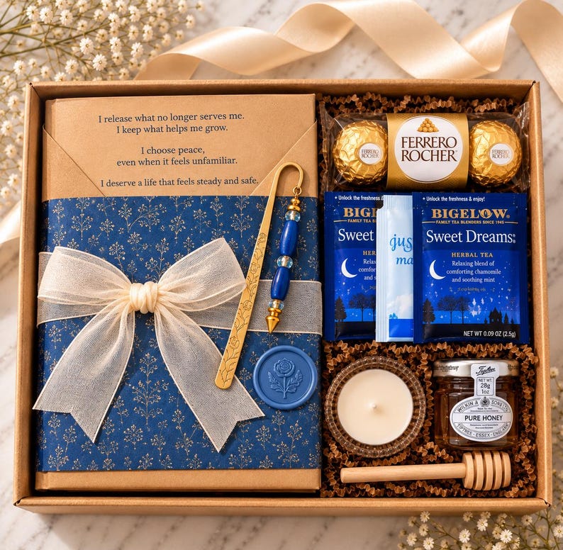 A gift box containing a blue-wrapped journal with a white ribbon, a gold-toned metal bookmark with blue beads, a gold pen, two Ferrero Rocher chocolates, two Bigelow Sweet Dreams tea bags, a small candle, and a jar of honey with a dipper.