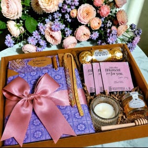 May include: A gift box containing a purple patterned notebook with a large dusty pink satin bow, a gold beaded bookmark, a Ferrero Rocher chocolate, three tea bags labeled Take this Moment just for you, a glass candle, and a small jar of honey.
