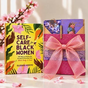 May include: A yellow book titled Self-Care for Black Women by Oludara Adeeyo with floral illustrations, next to a pink glittery gift box wrapped with a sheer pink ribbon, containing a gold pen and two purple cards with floral accents.