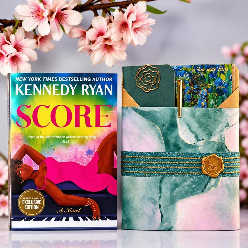 A book titled Score by Kennedy Ryan featuring a pink-clad figure on a piano, next to a wrapped gift set containing a green notebook with a rose-shaped paperclip, a gold pen, and a floral bookmark, all secured with teal ribbon and a gold wax seal.