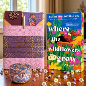 A book titled Where the Wildflowers Grow by Terah Shelton Harris next to a purple patterned gift box containing a notebook, a gold metal rose paperclip, and a floral bookmark, with a small round patterned tin in the foreground.