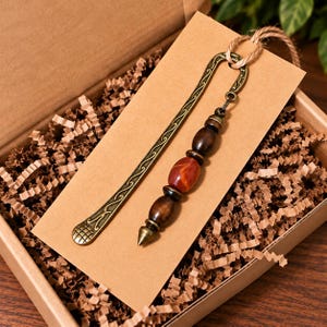 May include: A metal bookmark with an antique bronze finish featuring an intricate scroll pattern. Attached to the top is a dangling charm consisting of two dark brown wooden beads and one smooth, polished orange-red stone bead with a pointed tip.