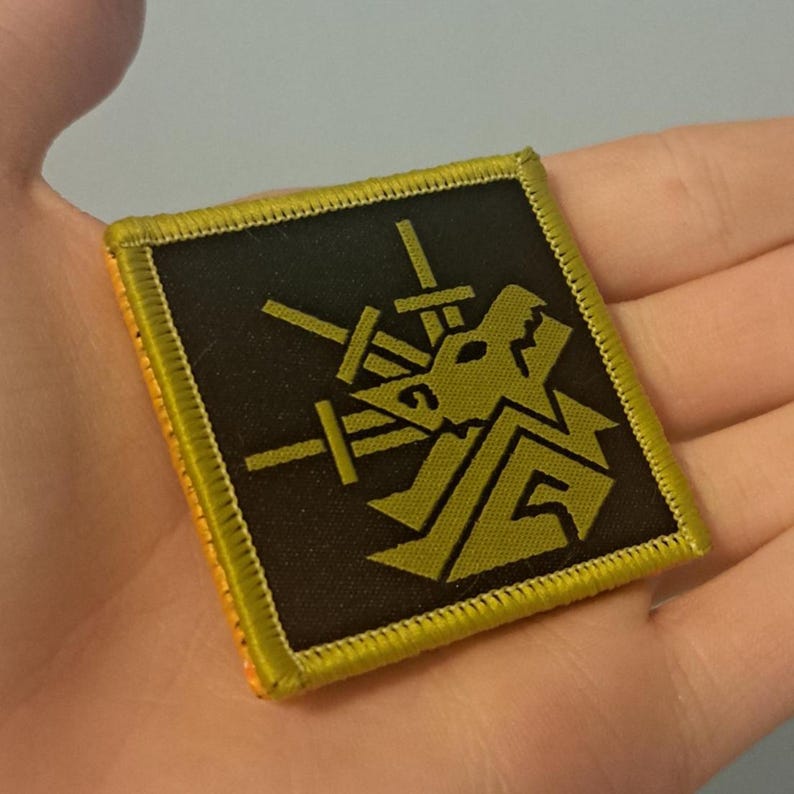 May include: Square patch with a black background and a gold-colored design featuring a stylized wolf head and crossed swords. The patch has a gold border and is likely an emblem or insignia.