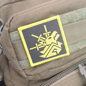 May include: A square patch with a yellow border and a black center, featuring a yellow graphic of a dragon and crossed swords. The patch is attached to a brown fabric surface, possibly a bag or backpack.