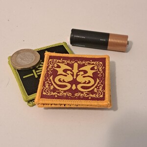 May include: A square, maroon patch with a gold dragon design and a gold border. A black and green patch with Japanese text and a coin are next to the patch. A standard AA battery is also in the image.