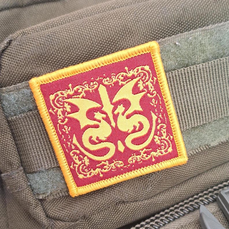 May include: Square patch with a red background and a gold dragon design. The patch has a gold border and is attached to a green fabric surface. The dragon design is stylized and symmetrical, with intricate details.