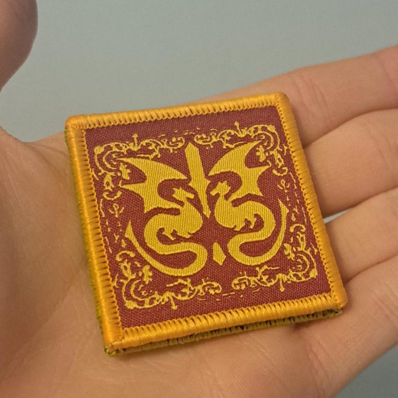 May include: Square patch with a red background and a gold border. The patch features a gold dragon design in the center, with ornate gold scrollwork around the edges. The patch is held in a hand.