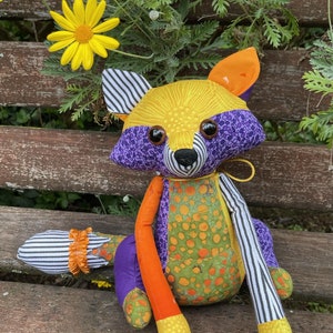 May include: A colourful, handmade raccoon doll with a patchwork design. The doll has a yellow head, purple accents, orange arms, and a green and orange patterned body. The tail is striped, and the doll has large, round eyes.