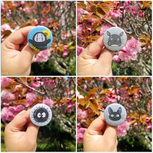 Könnte beinhalten: Vier verschiedene Anstecknadeln mit Figuren aus dem Animationsfilm 'Mein Nachbar Totoro'. Die Nadeln sind rund und haben einen weißen Hintergrund mit schwarzen Umrissen. Die Figuren sind Ohne Gesicht, Totoro, der Katzenbus und Jiji. Die Nadeln werden vor einem Hintergrund aus rosa Kirschblüten gezeigt.
