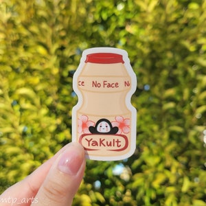 Peut inclure: Un autocollant repr&eacute;sentant une bouteille de Yakult blanche avec un bouchon rouge et des fleurs roses. La bouteille porte les inscriptions "No Face No" et "Yakult". Un personnage de dessin anim&eacute; avec un visage noir et des joues rouges se trouve &agrave; l'int&eacute;rieur de la bouteille.