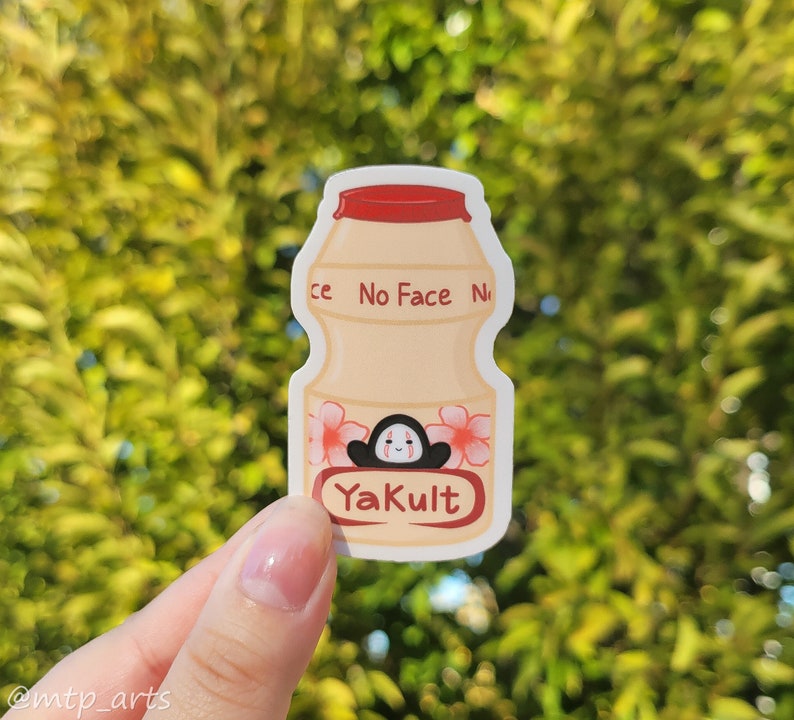 Peut inclure: Un autocollant repr&eacute;sentant une bouteille de Yakult blanche avec un bouchon rouge et des fleurs roses. La bouteille porte les inscriptions "No Face No" et "Yakult". Un personnage de dessin anim&eacute; avec un visage noir et des joues rouges se trouve &agrave; l'int&eacute;rieur de la bouteille.