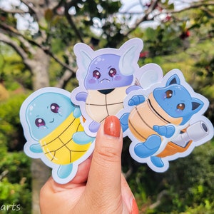 May include: Three cartoon turtle stickers with different expressions. The stickers are blue, purple, and yellow with brown shells. The stickers are on a hand against a green background.