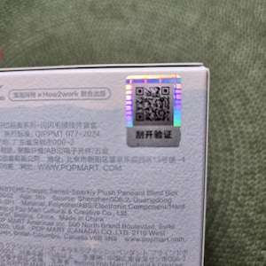 K&ouml;nnte beinhalten: Nahaufnahme einer wei&szlig;en Box mit Text und einem holografischen Aufkleber. Der Aufkleber hat einen QR-Code und chinesische Schriftzeichen. Die Box ist Teil der Monsters Classic Series.