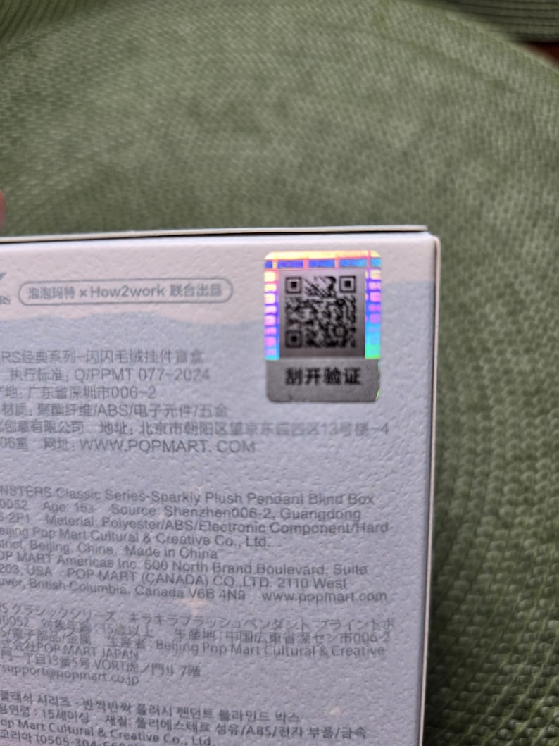 K&ouml;nnte beinhalten: Nahaufnahme einer wei&szlig;en Box mit Text und einem holografischen Aufkleber. Der Aufkleber hat einen QR-Code und chinesische Schriftzeichen. Die Box ist Teil der Monsters Classic Series.