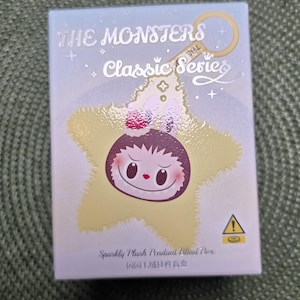 K&ouml;nnte beinhalten: Hellblaue Box mit dem Text "THE MONSTERS Classic Series" und einer sternf&ouml;rmigen Charakterillustration. Die Box hat auch den Text "Sparkly Plush Pendant Blind Box" und ein Warnsymbol.