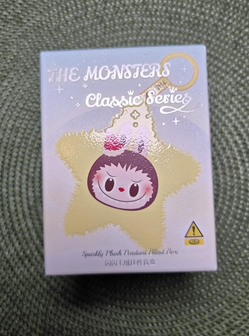K&ouml;nnte beinhalten: Hellblaue Box mit dem Text "THE MONSTERS Classic Series" und einer sternf&ouml;rmigen Charakterillustration. Die Box hat auch den Text "Sparkly Plush Pendant Blind Box" und ein Warnsymbol.