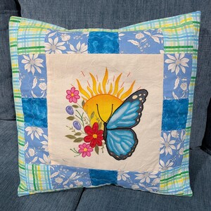 May include: A square throw pillow featuring a central cream-colored panel with an embroidered design of a yellow sun, a blue butterfly, and colorful flowers. The border consists of a patchwork pattern in shades of blue, white, green, and yellow.