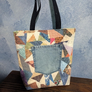 May include: A patchwork tote bag with a denim pocket. The bag features a variety of fabric patterns in shades of blue, brown, green, and pink. It has black handles and is designed for carrying items.