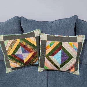 May include: Two square throw pillows featuring a central diamond pattern constructed from multi-colored fabric strips in shades of orange, green, purple, and blue. The pillows have a dark gray border and a light blue trim along the outer edges.