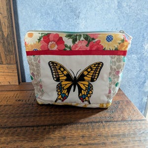 May include: A quilted fabric pouch featuring a central embroidered yellow and black butterfly on a white background. The top section displays a floral pattern with red, yellow, and pink flowers. A thin red ribbon trim separates the two sections.