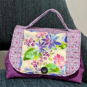 May include: A quilted fabric handbag featuring a purple and white floral print on the outer edges and a central panel with a colorful watercolor-style floral pattern in shades of blue, pink, and green, secured with a single dark button.