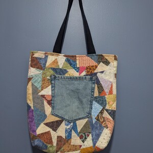 May include: A tote bag constructed from a patchwork of various multicolored fabric scraps featuring diverse patterns. The bag includes a central blue denim pocket and two black fabric shoulder straps, showcasing a quilted texture throughout the design.