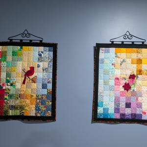May include: Two rectangular patchwork wall quilts hanging from black metal hangers. The left quilt features a red cardinal bird on a branch amidst a grid of multicolored fabric squares. The right quilt displays a floral motif on a gradient grid.