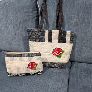 May include: A quilted tote bag and a matching zippered pouch, both featuring a patchwork design of black, white, and gray fabrics with a prominent red rose appliqué with green leaves on the front of each item. The tote has two fabric handles.