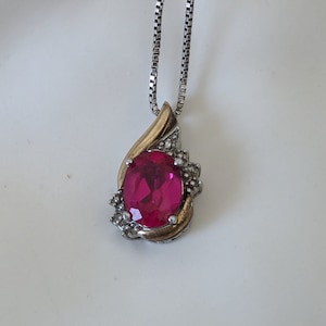 May include: A silver chain necklace with a pink oval gemstone pendant set in gold. The pendant is surrounded by smaller clear gemstones.